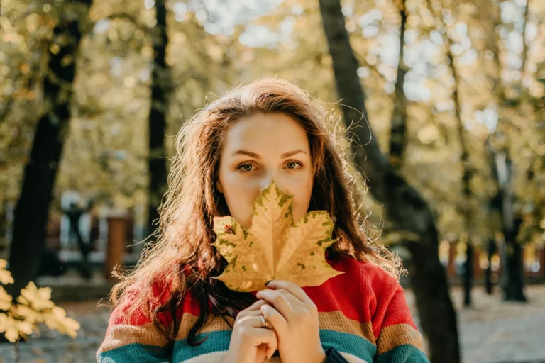 Una ragazza ha una foglia autunnale in mano e nasconde parte del viso coprendo la bocca.